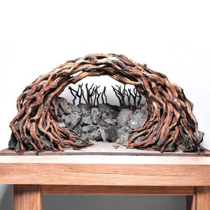 May include: A handcrafted sculpture of a natural archway. The arch is formed by intertwined, textured brown roots, spanning over a stone wall. Inside the arch, there are stylized, dark tree silhouettes. The sculpture sits on a wooden base.