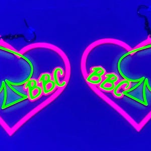May include: Pair of neon pink heart-shaped earrings with a green spade design and the letters "BBC" in green. The earrings have black ear wires and are set against a dark blue background.