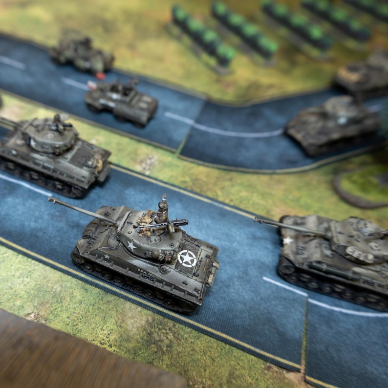 May include: Miniature military tanks in various shades of green and gray are positioned on a blue road, part of a tabletop game. The tanks feature detailed designs, including star insignias and gun turrets. The scene suggests a strategic war game.