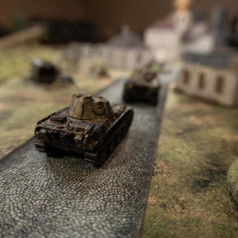 May include: A detailed miniature military tank model in green and brown hues, positioned on a grey road. The tank is the primary focus, with a blurred background suggesting a battlefield scene. The model has intricate features.