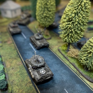 May include: Miniature military tanks on a model road with trees. The tanks are painted in shades of gray and green, with a white star emblem on the front. The road is gray with white lane markings, and the scene is set against a backdrop of green terrain and miniature trees.