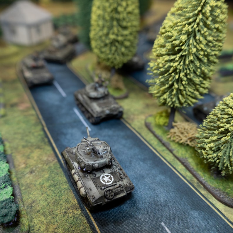 May include: Miniature military tanks on a model road with trees. The tanks are painted in shades of gray and green, with a white star emblem on the front. The road is gray with white lane markings, and the scene is set against a backdrop of green terrain and miniature trees.