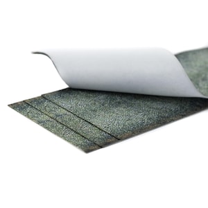 May include: A close-up of a stack of textured, dark green material with a white backing. The top layer is partially peeled back, revealing the underside. The material has a mottled, leather-like appearance.