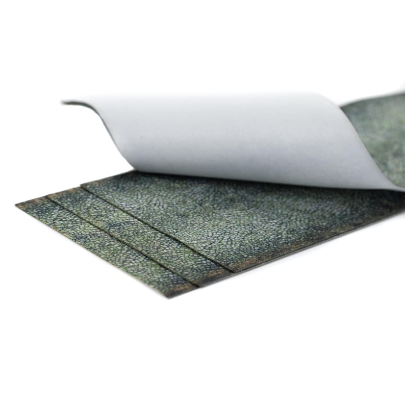May include: A close-up of a stack of textured, dark green material with a white backing. The top layer is partially peeled back, revealing the underside. The material has a mottled, leather-like appearance.