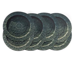 Puede incluir: Un conjunto de seis posavasos redondos con apariencia de piedra. Cada posavasos presenta un diseño circular detallado con un emblema central y un borde exterior de patrones repetidos en tonos grises y verdes. Los posavasos están dispuestos en un patrón ligeramente superpuesto.