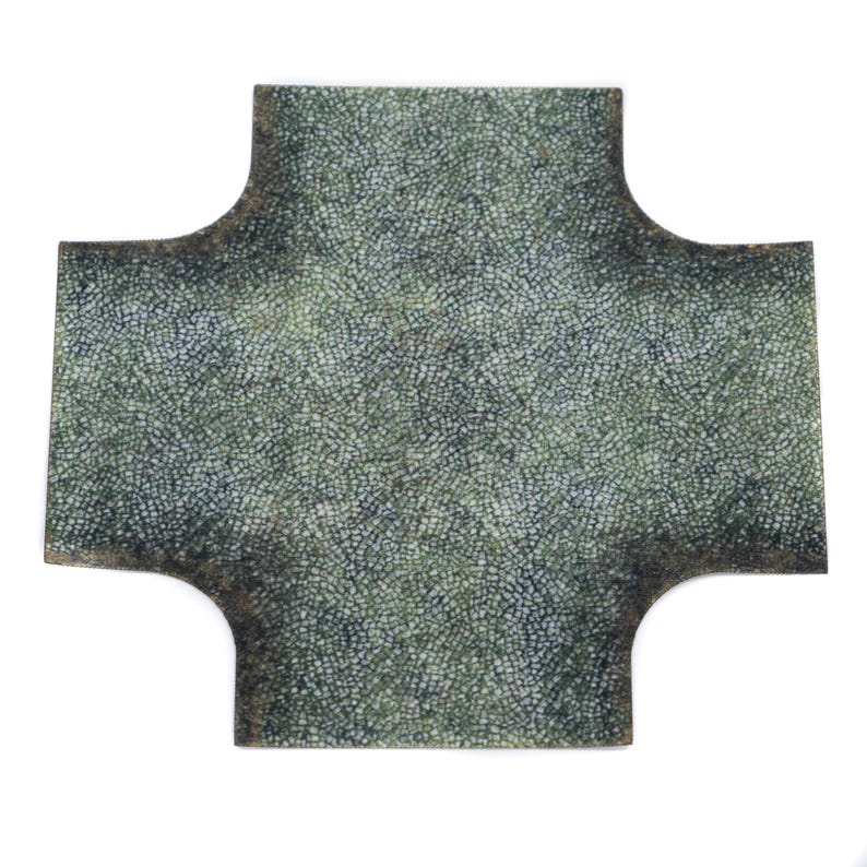 May include: A cross-shaped tile with a mosaic design in green and grey hues. The edges have a darker, aged look. The tile is set against a white background.