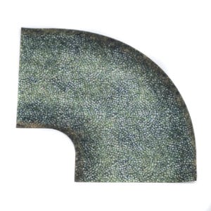 May include: A quarter-circle mat with a mosaic pattern in green and grey. It has a rounded corner and a curved cutout. The edges have a darker, weathered appearance. The mat's texture resembles small, closely packed stones.