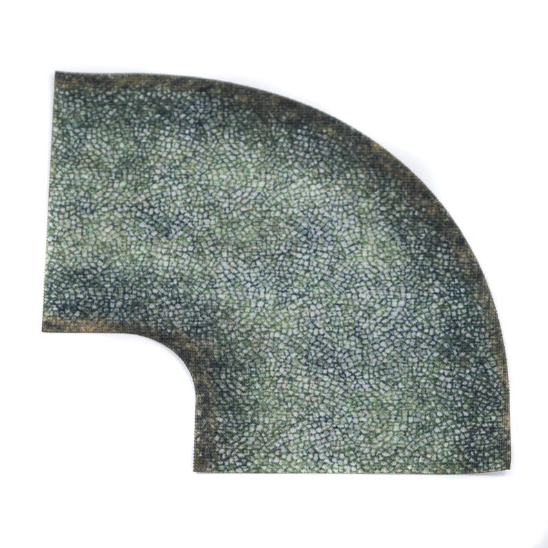 May include: A quarter-circle mat with a mosaic pattern in green and grey. It has a rounded corner and a curved cutout. The edges have a darker, weathered appearance. The mat's texture resembles small, closely packed stones.