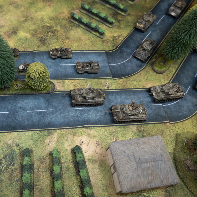 May include: Miniature military tanks on a model road with trees and a building. The tanks are painted in shades of green and brown, and the road is a dark blue-gray color. The scene is set on a green and brown terrain.