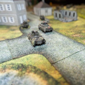 May include: A collection of miniature military vehicles on a cobblestone road, with model buildings in the background. The vehicles are camouflaged, and the road is grey. The scene suggests a tabletop war game or diorama.