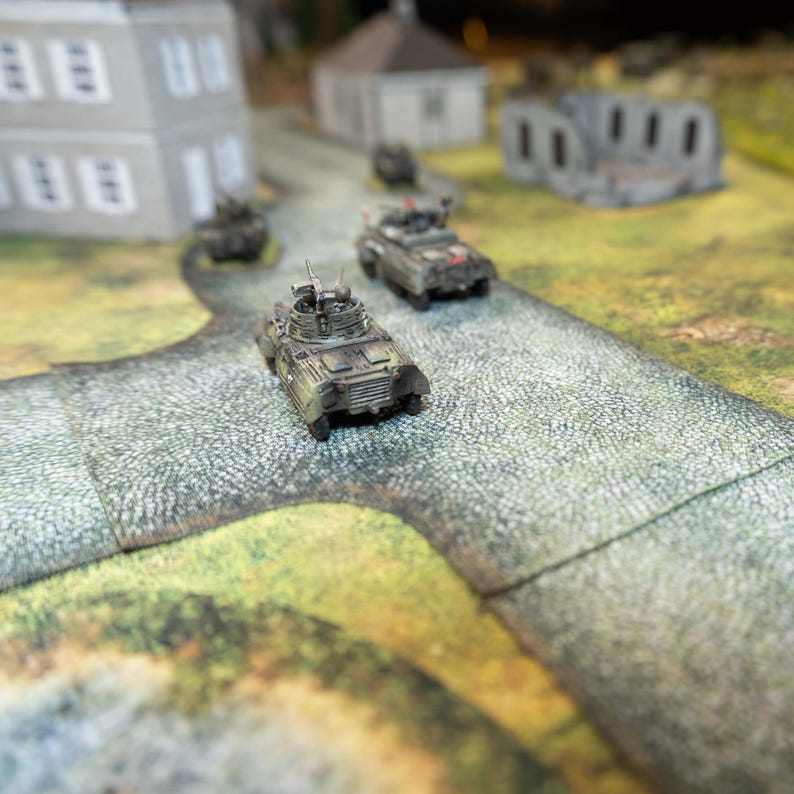 May include: A collection of miniature military vehicles on a cobblestone road, with model buildings in the background. The vehicles are camouflaged, and the road is grey. The scene suggests a tabletop war game or diorama.