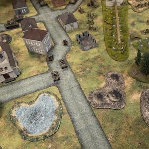 May include: A detailed tabletop war game setup featuring miniature tanks, buildings, and terrain elements. The scene includes roads, a pond, trees, and structures, creating a battlefield environment. The game board is green and grey.