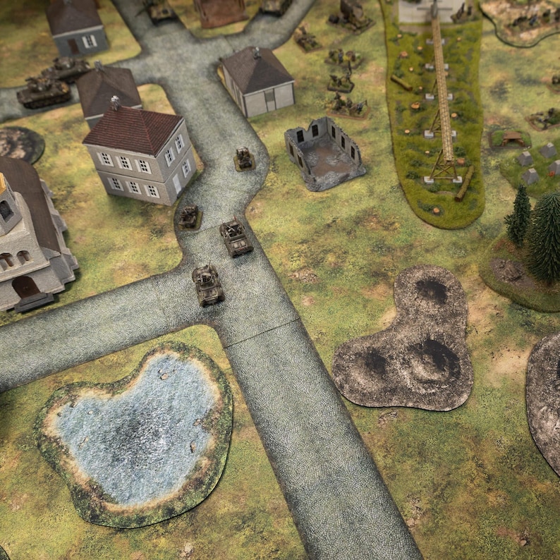 May include: A detailed tabletop war game setup featuring miniature tanks, buildings, and terrain elements. The scene includes roads, a pond, trees, and structures, creating a battlefield environment. The game board is green and grey.