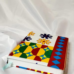 Könnte beinhalten: Ein Notizbuch mit festem Einband und farbenfrohem Muster. Das Design zeigt geometrische Formen in Rot, Grün, Gelb und Blau auf weißem Hintergrund. Ein hellblaues Lesezeichenband ist sichtbar.