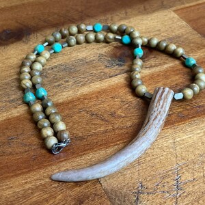 May include: A brown wooden beaded necklace with turquoise and silver accents. The necklace has a long, curved antler pendant.
