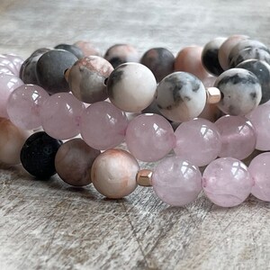 May include: Three stacked bracelets with pink and gray gemstone beads. The top bracelet has gray and white marbled beads, the middle bracelet has pink and gray marbled beads, and the bottom bracelet has light pink beads.
