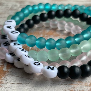 May include: Four beaded bracelets with white letter beads spelling out "I LOVE YOU" and "MARSO" and "GIRL" and "N" on a wooden surface.