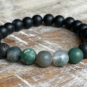 May include: A beaded bracelet featuring black matte onyx beads, porous black lava beads, and a mix of green and gray patterned stone beads. The bracelet is strung on a stretch cord and rests on a wooden surface.