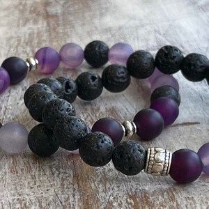 May include: Two bracelets with purple and black beads. One bracelet has dark purple beads and black lava rock beads. The other bracelet has light purple and dark purple beads with black lava rock beads. Both bracelets have silver metal accents.