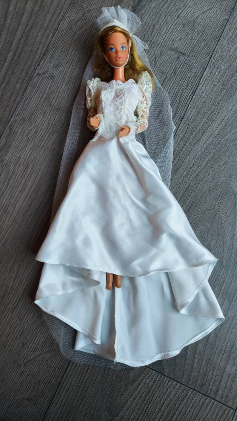 May include: A white bridal gown with a lace bodice and a tulle veil. The doll is wearing a white headband with a flower.