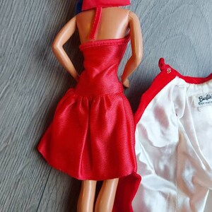 May include: A red dress with a ruffled hem and a matching red cape. The dress is made of a shiny fabric and has a halter neckline. The cape has a white lining and a button closure.