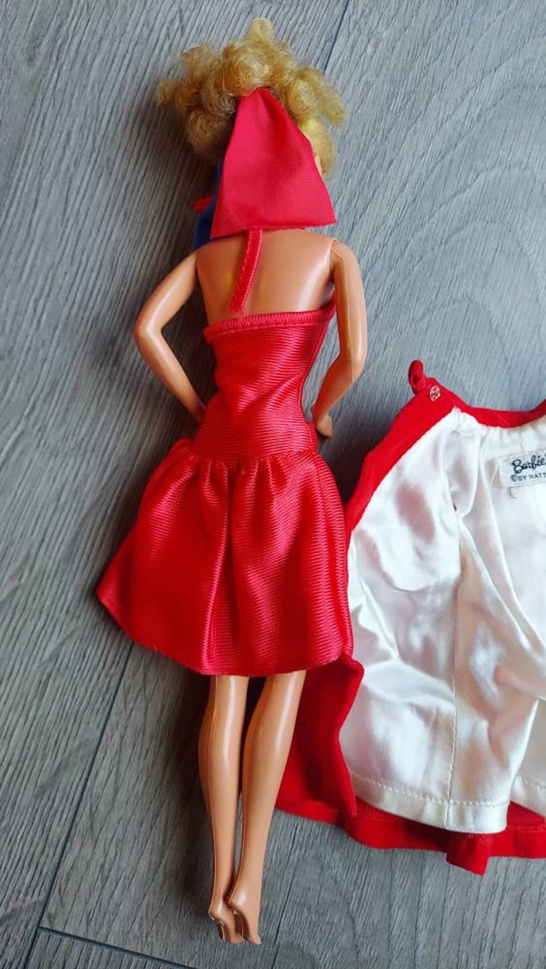 May include: A red dress with a ruffled hem and a matching red cape. The dress is made of a shiny fabric and has a halter neckline. The cape has a white lining and a button closure.