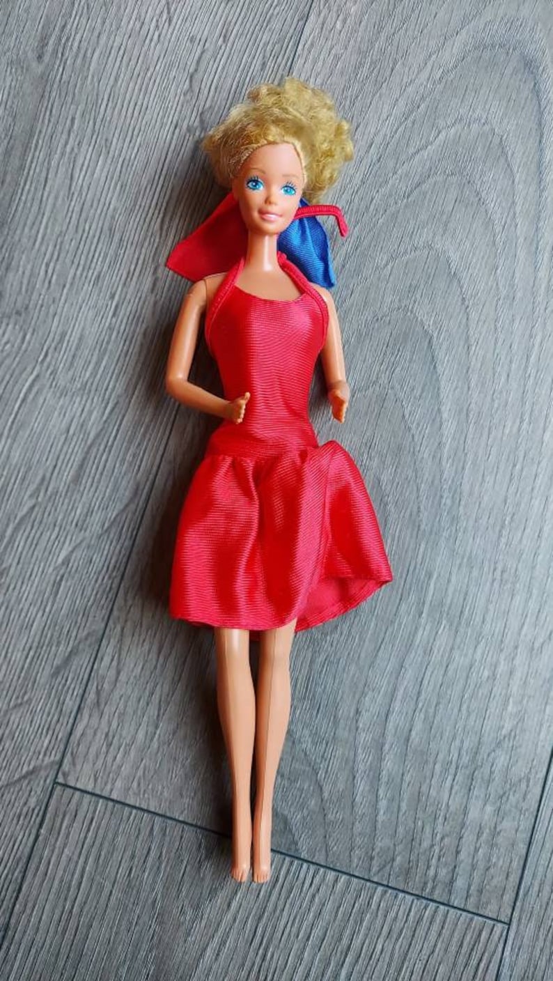 May include: A vintage Barbie doll wearing a red satin dress with a blue bow. The doll has blonde hair and blue eyes.
