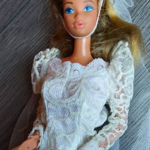 May include: A white bridal doll with blonde hair, wearing a white lace dress and a white veil. The doll has blue eyes and a pink blush.