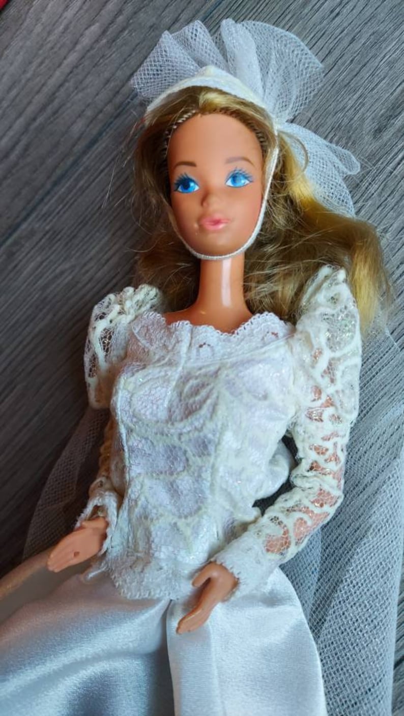 May include: A white bridal doll with blonde hair, wearing a white lace dress and a white veil. The doll has blue eyes and a pink blush.