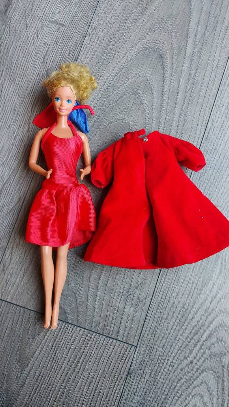 May include: A vintage Barbie doll wearing a red dress and a blue ribbon. The doll is also wearing a red velvet coat.