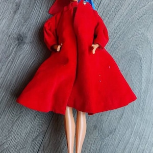 May include: A vintage Barbie doll wearing a red coat with a blue and red bow. The doll has blonde hair and blue eyes.