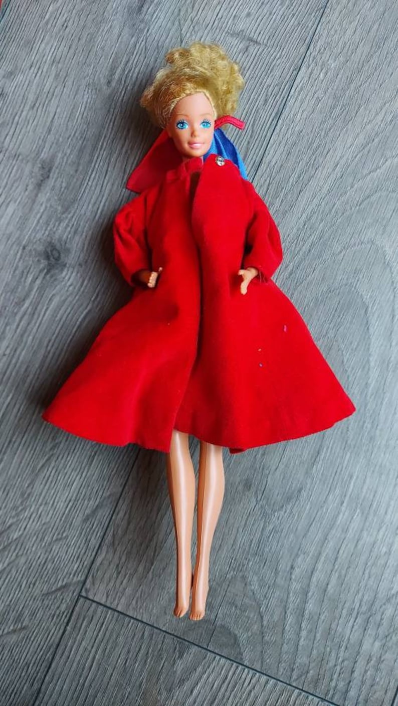 May include: A vintage Barbie doll wearing a red coat with a blue and red bow. The doll has blonde hair and blue eyes.