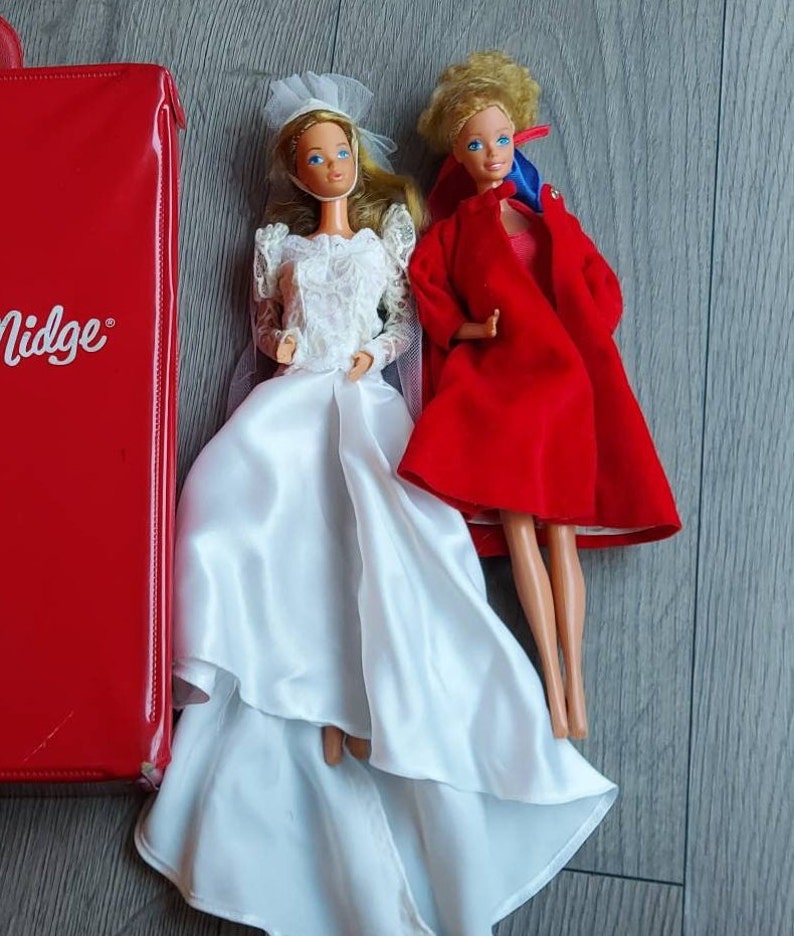 May include: Two Barbie dolls, one wearing a white wedding dress and the other wearing a red coat. The dolls are standing on a wooden surface.