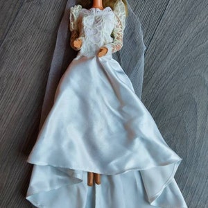 May include: A white bridal doll with a lace bodice and a flowing satin skirt. The doll is wearing a white veil and has blonde hair and blue eyes.