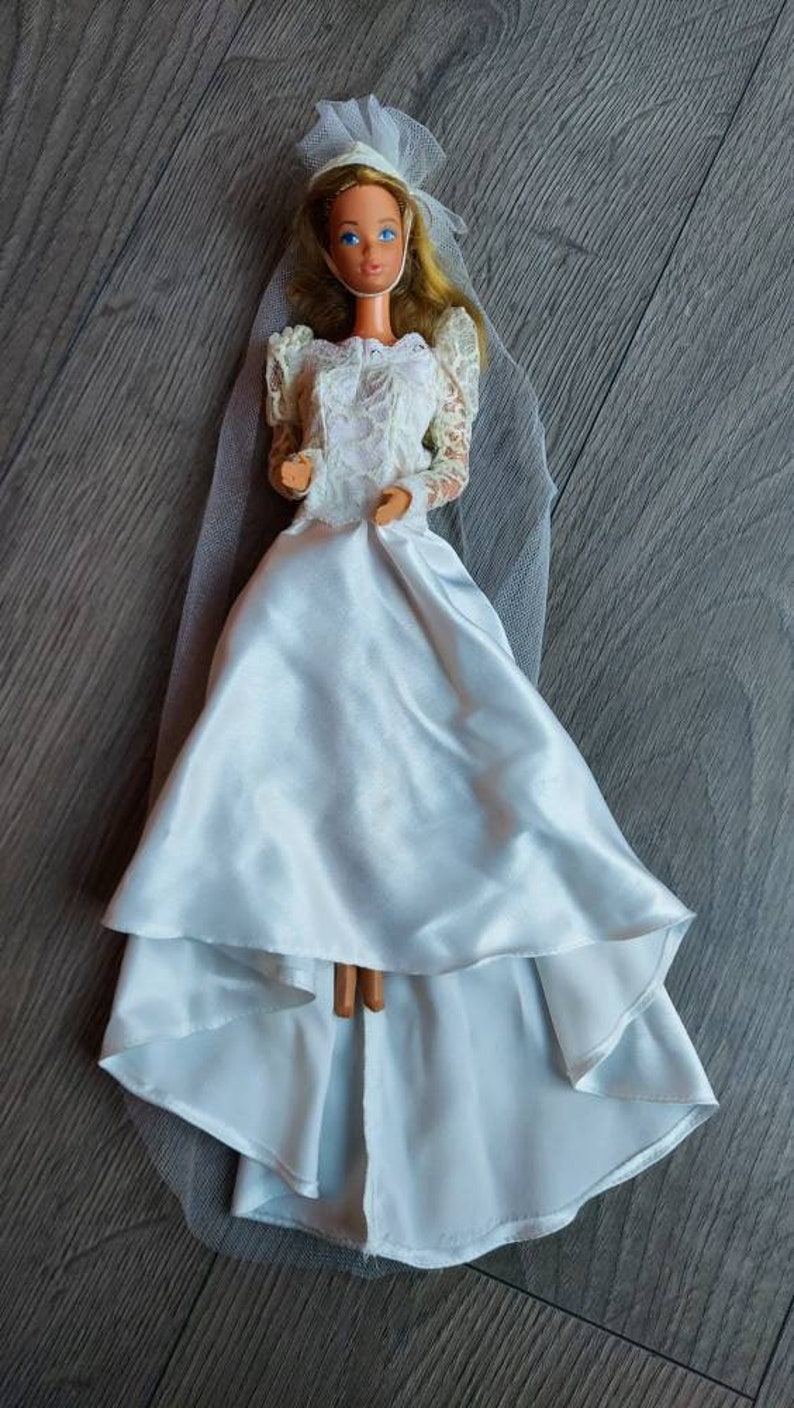 May include: A white bridal doll with a lace bodice and a flowing satin skirt. The doll is wearing a white veil and has blonde hair and blue eyes.