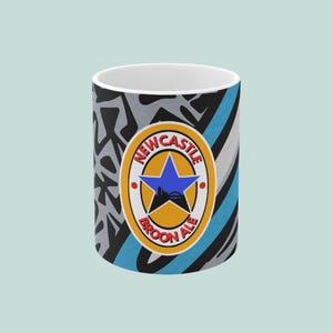 May include: A white ceramic mug with a colorful design. The mug features the Newcastle Brown Ale logo with a blue star and a cityscape silhouette. The background has a mix of black, gray, and blue patterns.