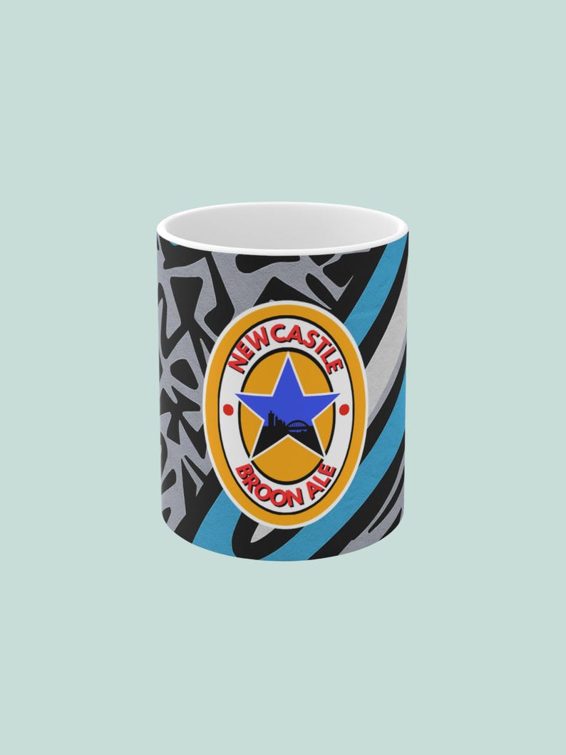 May include: A white ceramic mug with a colorful design. The mug features the Newcastle Brown Ale logo with a blue star and a cityscape silhouette. The background has a mix of black, gray, and blue patterns.