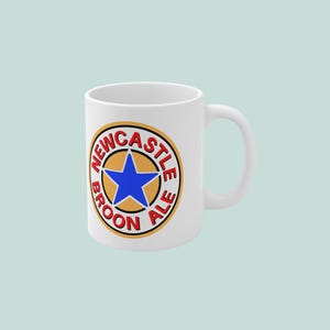 Puede incluir: Taza de cerámica blanca con el logotipo de Newcastle Brown Ale. El logotipo es una estrella azul dentro de un círculo dorado, con las palabras "Newcastle Brown Ale" en rojo. La taza tiene un asa curva.