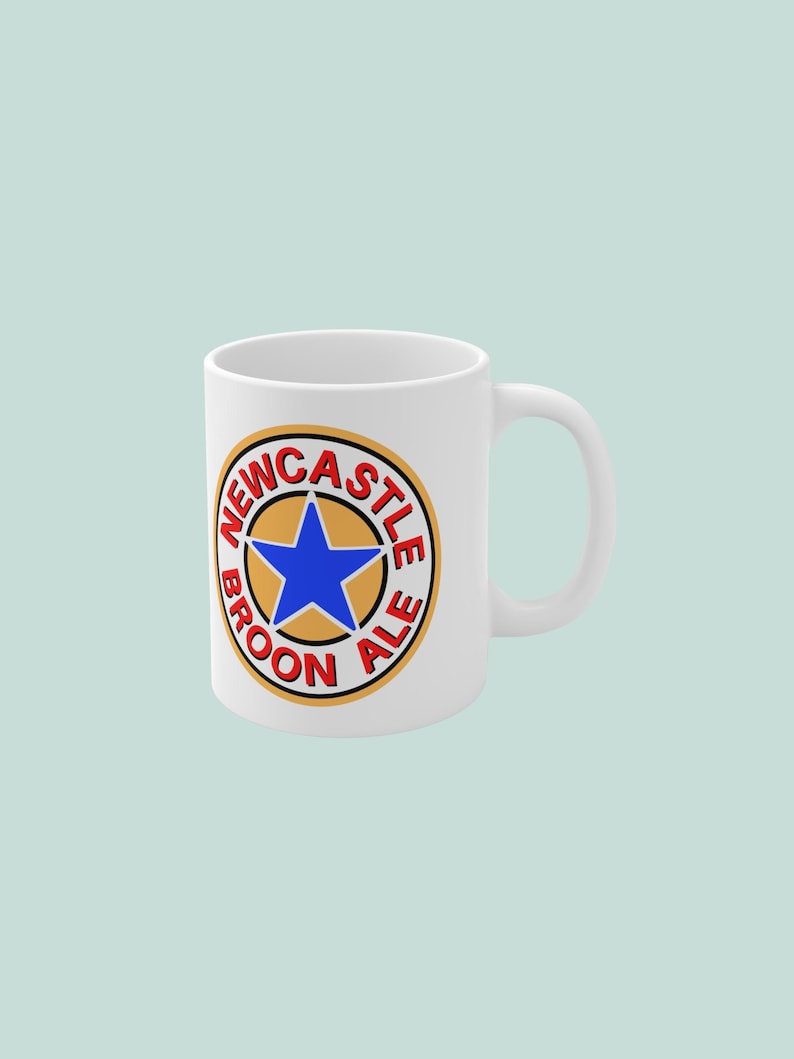 K&ouml;nnte beinhalten: Wei&szlig;e Keramiktasse mit dem Logo von Newcastle Brown Ale. Das Logo zeigt einen blauen Stern in einem goldenen Kreis mit den Worten "Newcastle Brown Ale" in Rot. Die Tasse hat einen gebogenen Griff.