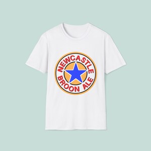 Puede incluir: Camiseta blanca con un logotipo circular. El logotipo dice "NEWCASTLE BROON ALE" en rojo, naranja y negro, con una estrella azul en el centro. La camiseta tiene cuello redondo y mangas cortas.