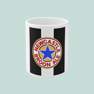 Puede incluir: Taza de cerámica blanca con rayas verticales negras y un emblema circular. El emblema presenta una estrella azul en un círculo dorado, con las palabras "NEWCASTLE BROON ALE" en letras rojas. La taza es cilíndrica.