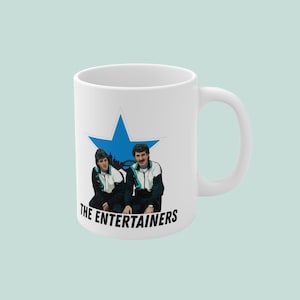 May include: White ceramic mug featuring a blue star with an illustration of two people in tracksuits. The text "THE ENTERTAINERS" is printed below the illustration. The mug has a white handle.