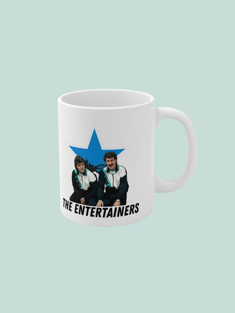 May include: White ceramic mug featuring a blue star with an illustration of two people in tracksuits. The text "THE ENTERTAINERS" is printed below the illustration. The mug has a white handle.