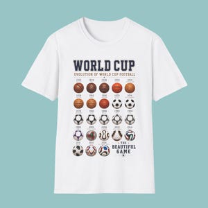 May include: A white short-sleeve t-shirt featuring a graphic print titled WORLD CUP EVOLUTION OF WORLD CUP FOOTBALL. The design displays a chronological grid of various footballs used from 1930 to 2026, ending with the text THE BEAUTIFUL GAME.