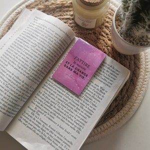 Peut inclure: Un marque-page rose et violet avec le texte "J'attire le succès et la chance dans ma vie" en lettres blanches. Le marque-page est sur un livre avec un fond blanc.