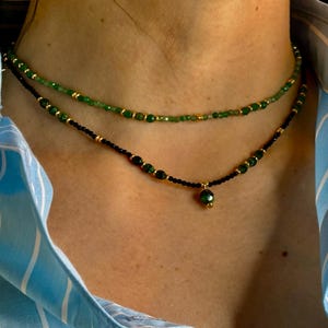 May include: A layered necklace with black and green beads. The top layer is made of green beads and the bottom layer is made of black beads. A single green gemstone pendant hangs from the bottom layer.