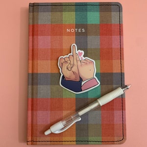 May include: A notebook with a plaid cover in shades of orange, green, and brown, with the word "NOTES" printed on the front. A sticker of two hands with a heart and a white pen are on the notebook.