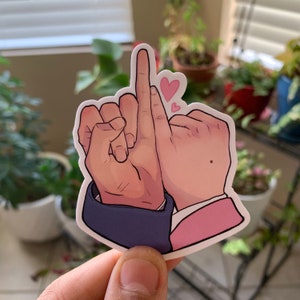 May include: A sticker featuring two hands with intertwined fingers, one hand giving the middle finger. The hands are depicted with a cartoon style, with pink hearts floating above. The sleeve cuffs are blue and pink.