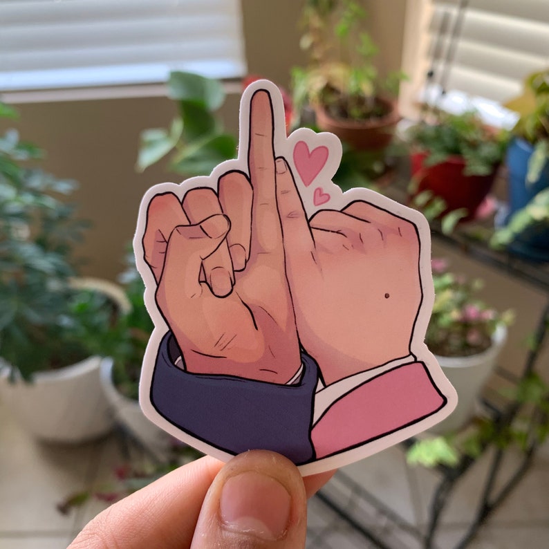 May include: A sticker featuring two hands with intertwined fingers, one hand giving the middle finger. The hands are depicted with a cartoon style, with pink hearts floating above. The sleeve cuffs are blue and pink.