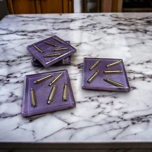 May include: Four square purple coasters with gold bullet casings embedded in the resin. The coasters are on a white marble surface.